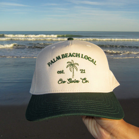 Cap with 'Palm Beach Local' text held on a beach with ocean waves in the background