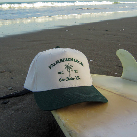 Cap with 'Palm Beach Local' text on a beach with a surfboard
